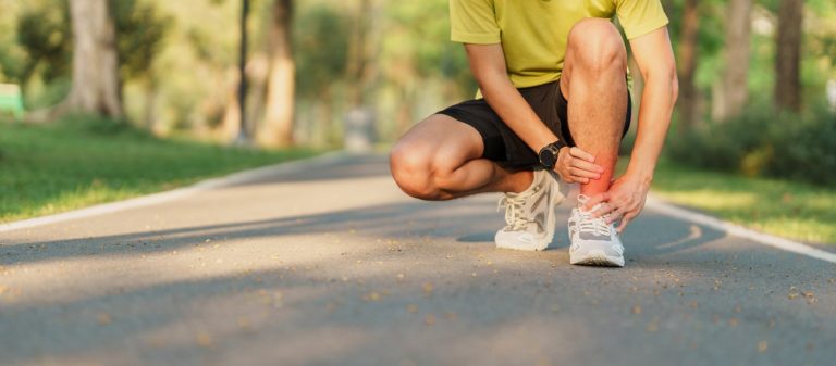 Runner on a pathway in wooded park kneeling and feeling his ankle to check for an ankle sprain